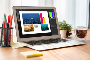 Laptop screen displaying a colorful logo design project with pencils, coffee, and a plant on a desk.